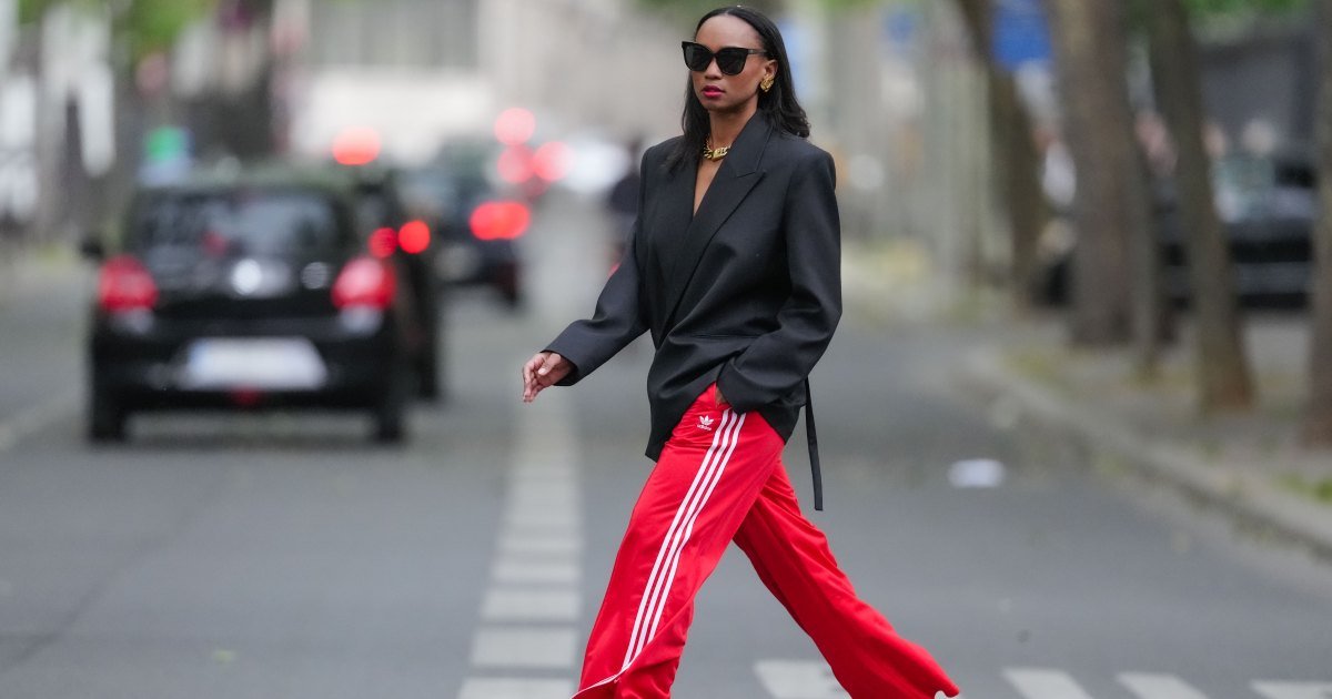 Emilie Joseph usa gafas de sol, aretes, lápiz labial rojo, blazer negro extragrande/estructurado de mezcla de lana virgen con cuello de solapa de Victoria Beckham para Mango, pantalones de chándal/pantalones deportivos extragrandes Adidas Classics en rojo a rayas, collar/cadena de oro grueso de Merbabe Grecia, zapatos: zapatos de tacón Newton de satén puntiagudo negro de Saint Laurent, durante una sesión de fotos de moda de estilo urbano, el 12 de mayo de 2024 en París, Francia.