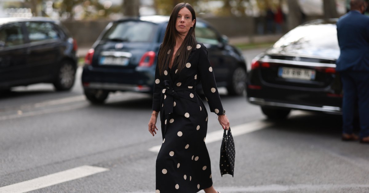 Una invitada a la Semana de la Moda luce aretes redondos de diamantes, un vestido midi negro de manga larga con estampado de lunares blancos, solapas de muesca y cinturón de corbata; una pulsera de oro; un reloj de oro rosa y negro de Rolex; un anillo de oro; un bolso rectangular negro con estampado de lunares blancos; y zapatos de tacón de punta negra, también con un patrón de lunares blancos antes del desfile Elie Saab Womenswear Primavera/Verano 2025 como parte de la Semana de la Moda de París el 28 de septiembre de 2024 en París, Francia.